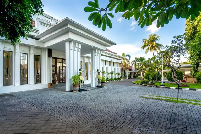 A white columned hotel entrance with a porte cochere stands over a cobblestone driveway, with palm trees, frangipani and statues beside the doorway.