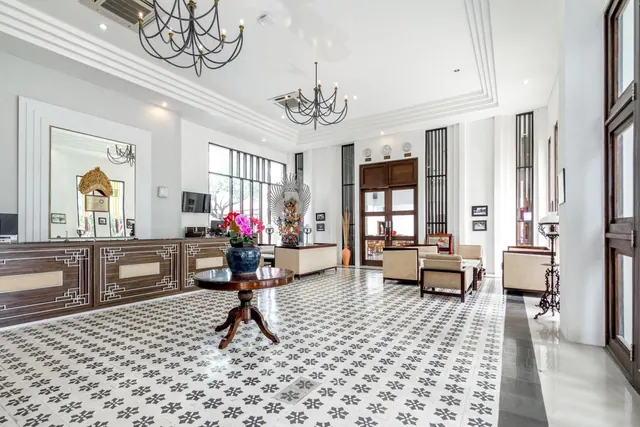 A hotel lobby with a reception desk, patterned tile floor, round wooden table with a flower vase, seating areas and two black chandeliers.