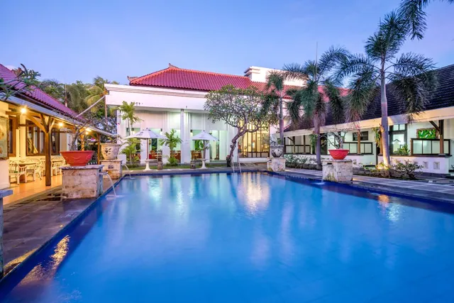 A large blue swimming pool at dusk with palm trees, red-tiled roof buildings, and white umbrellas on a terrace behind it.