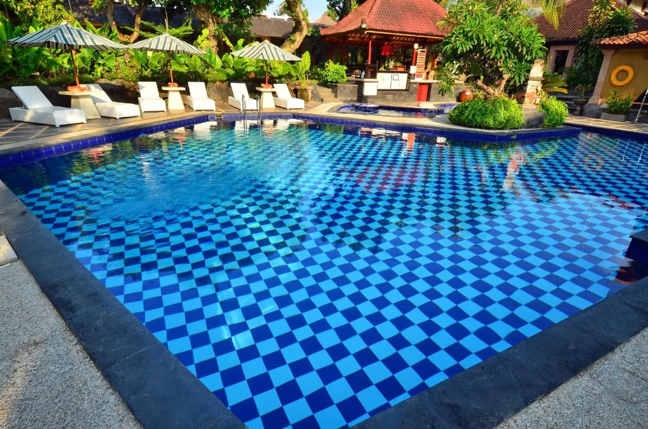 An outdoor swimming pool with blue checkered tiles, white sun loungers and striped umbrellas beside it, with a small pool and a red-roofed pavilion behind.