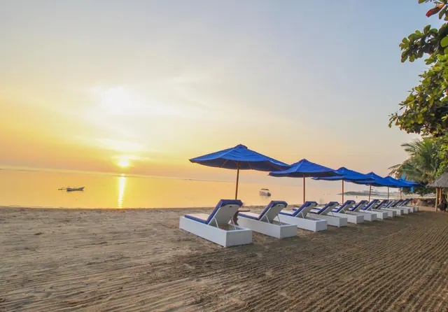 Rows of white sun loungers with blue cushions and blue parasols stand on a sandy beach facing the sea as the sun sets low over the water.