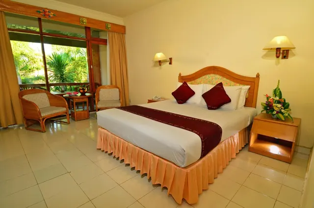 A hotel bedroom with a king bed, red cushions, wicker armchairs and a small table by a window looking onto palm trees.