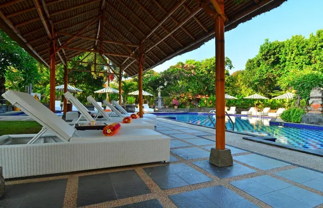 White wicker sun loungers with flower decorations sit under a thatched pavilion beside a blue-tiled swimming pool.