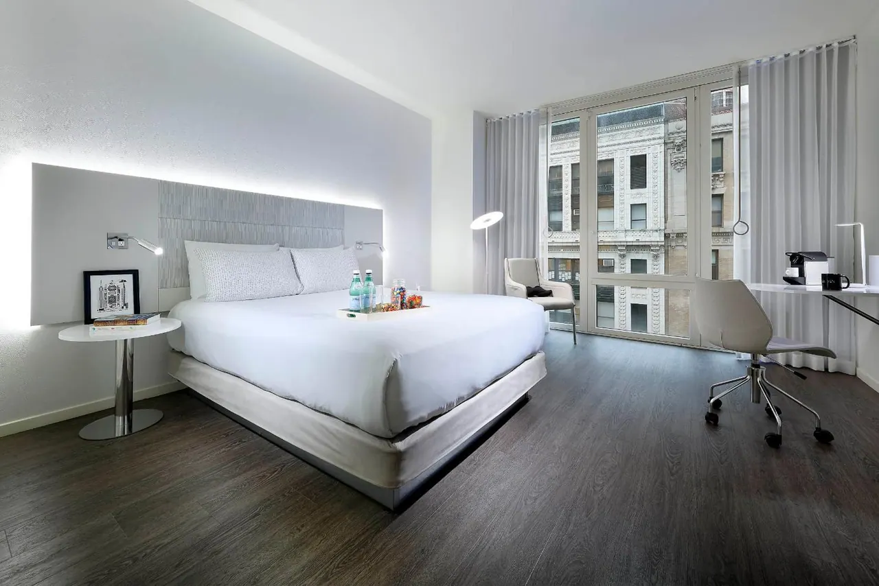 A king bed with white linens sits in a bedroom with a work desk, an armchair and floor-to-ceiling windows overlooking a building facade.
