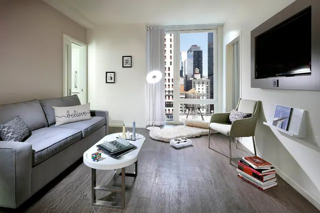 A living room with a grey sofa, a white oval coffee table, a wall-mounted television and a floor-to-ceiling window looking onto city high-rises.