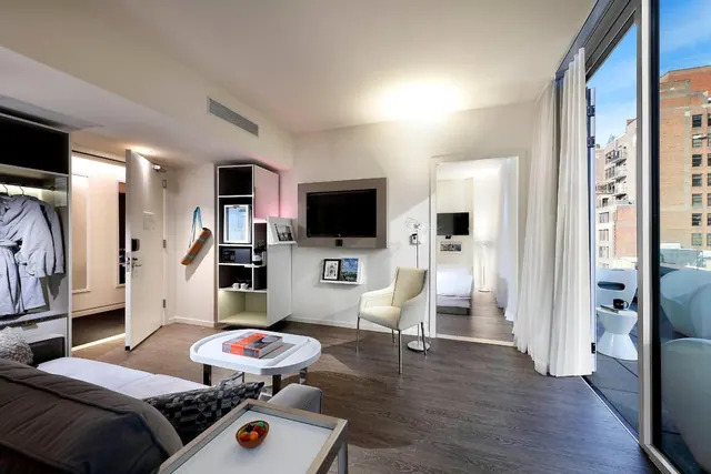 A modern hotel suite living area with a sofa, wall-mounted television and coffee table, with a glass door opening onto a balcony overlooking brick buildings.