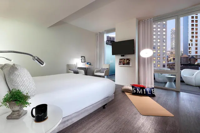 A hotel bedroom with a king bed, a seating area, a wall-mounted television and floor-to-ceiling windows opening onto a balcony with city buildings beyond.
