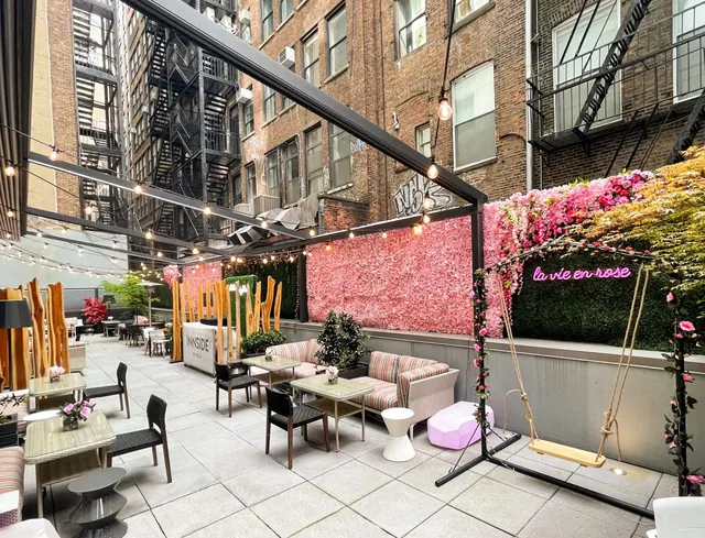 An outdoor terrace with low tables, cushioned sofas, a swing, a pink flower wall and a neon sign reading la vie en rose, surrounded by brick buildings and fire escapes.