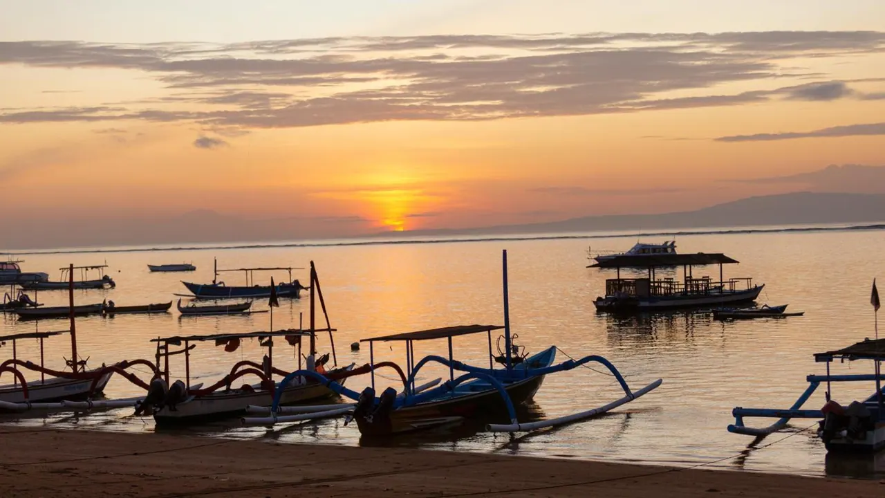 Outrigger boats are moored on a sandy beach while the low sun glows over calm water and distant hills.