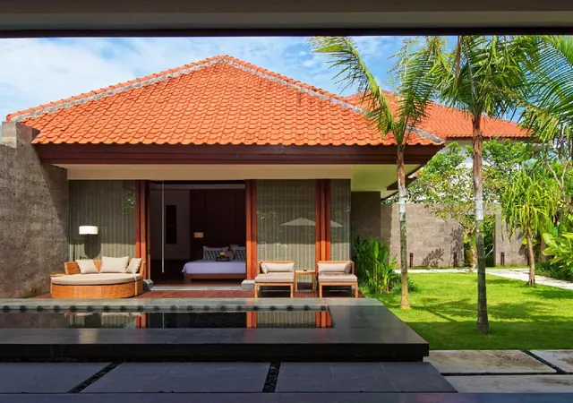 A single-storey villa with a terracotta tiled roof, open sliding doors showing a bedroom with a double bed, a rattan daybed and lounge chairs on the terrace, a dark pool in the foreground, and palm trees on the lawn beside it.