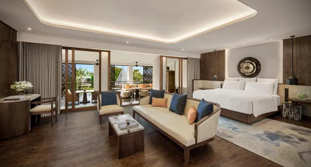A staged suite with a king bed, a cream sofa with cushions, an armchair, and glass doors opening onto a balcony with palm trees beyond.