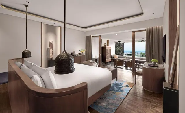 A bedroom with a large white bed, pendant lamps and a wooden bench at the foot, with glass doors leading to a seating area and a balcony overlooking the sea.