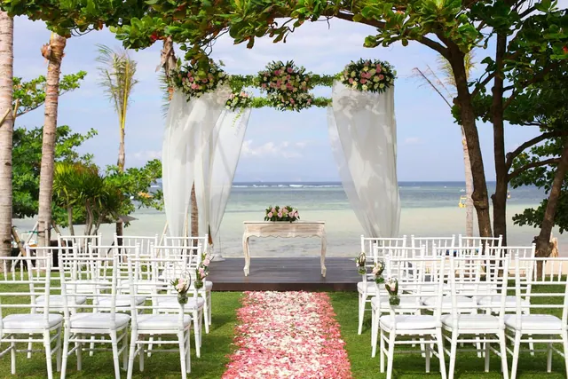 An outdoor wedding ceremony is set up on a lawn with white chairs, a floral arch with draped white fabric, a petal-strewn aisle and the sea visible behind.