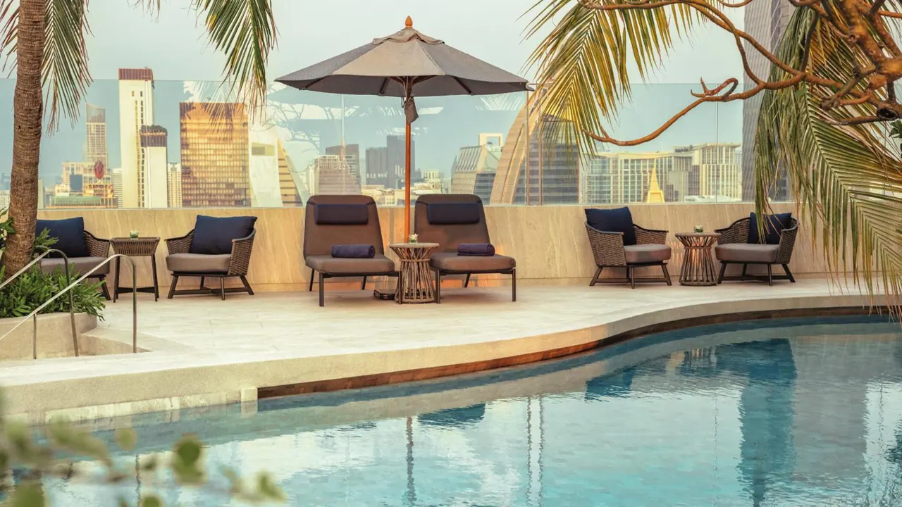 A pool deck with wicker lounge chairs and a shaded umbrella, with a city skyline visible beyond the glass railing.