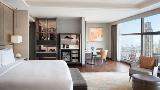 A hotel room with a king bed, a wardrobe and minibar unit, a round table with chairs, and floor-to-ceiling windows overlooking city high-rises.