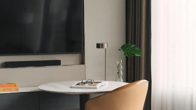 A round marble table holding a magazine, glasses and a small plant sits beside a wall-mounted television and a tan leather chair.