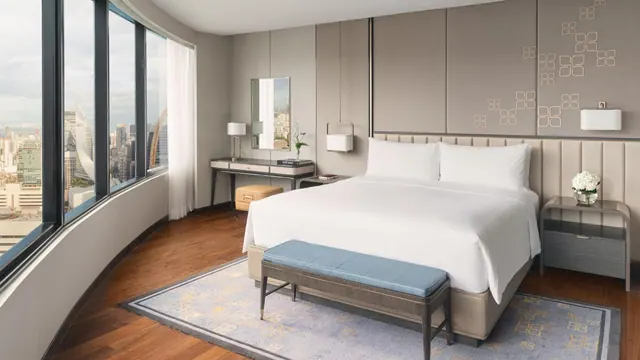 A bedroom with a made king bed, a blue upholstered bench at its foot, and curved floor-to-ceiling windows looking out over a city skyline.