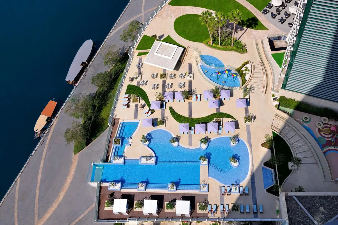 Aerial view of a hotel pool deck with a large freeform pool, a smaller pool, rows of sun loungers, purple shade umbrellas and a waterway along the left edge.