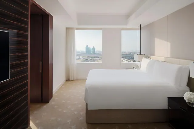 A bedroom with a made king bed, a dark wood-veneer wall with a wall-mounted television and a wardrobe, and two windows looking out over a city with a tower and a road.