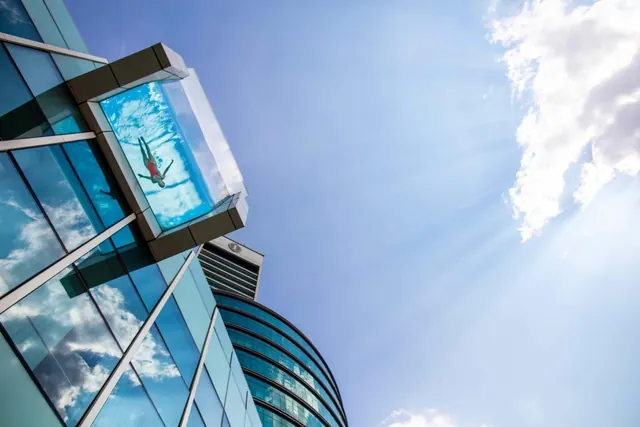 Low-angle view of a glass-walled building with a transparent cube pool projecting from its facade, with one person in the pool against a blue sky.