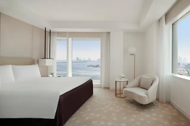 A bedroom with a king bed and an armchair beside floor-to-ceiling windows looking out over water toward a city skyline.