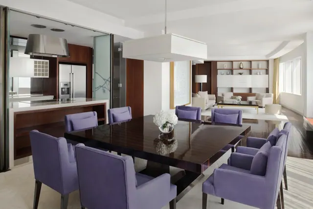 A dining area with a dark square table and lilac upholstered chairs, with a kitchen to the left and a sitting area with sofas to the right.