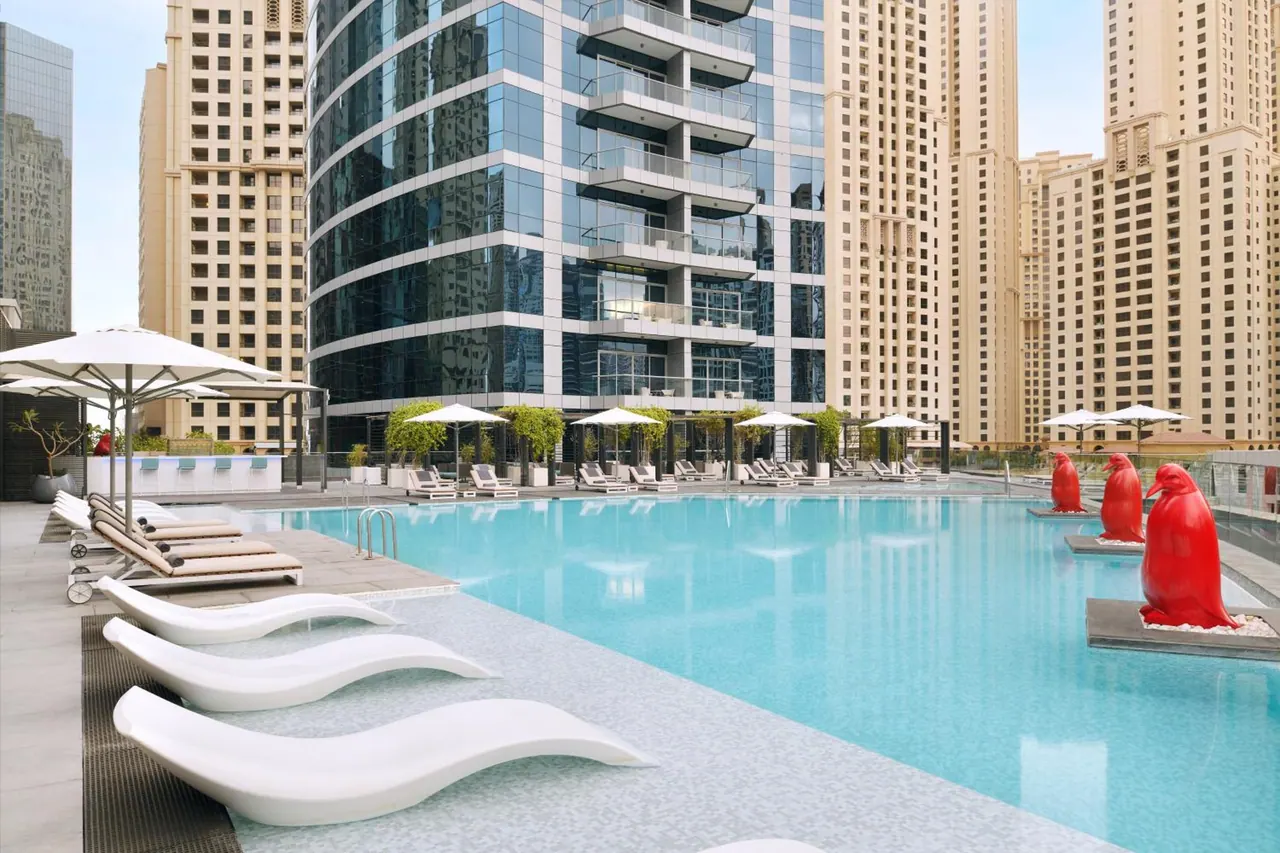 An outdoor swimming pool with white lounge chairs, shade umbrellas and red penguin sculptures in the water, surrounded by high-rise towers.