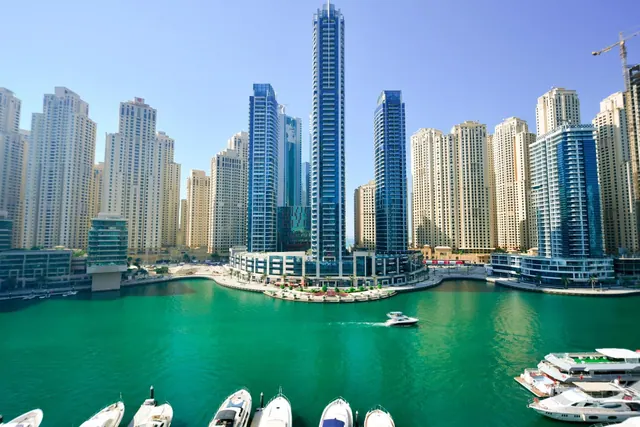 High-rise towers and moored yachts line a green marina waterway under a clear blue sky, seen from an elevated position.