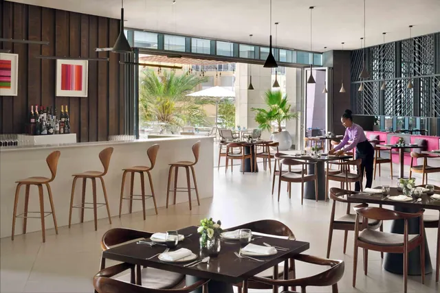 A restaurant dining room with wooden bar stools at a counter, set tables, pink banquette seating, and a staff member setting a table near large windows that look out onto palm trees.