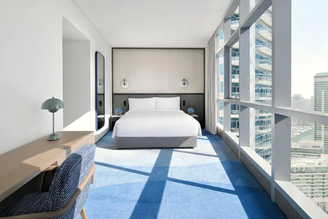 A bedroom with a king bed, blue carpet, a work desk, and floor-to-ceiling windows showing high-rise towers outside.