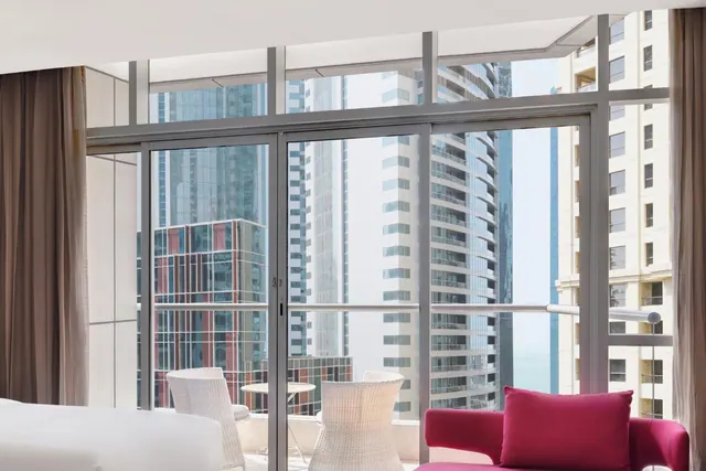 A hotel room with floor-to-ceiling glass doors opening onto a balcony with wicker chairs and a small table, overlooking high-rise towers, with a pink sofa and the edge of a white bed in the foreground.