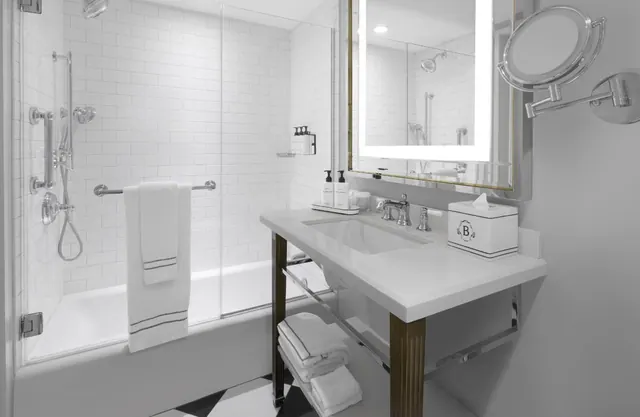 A white bathroom with a tub and glass-screened shower, a single undermount sink on a brass-legged console, a lit mirror and a magnifying mirror.
