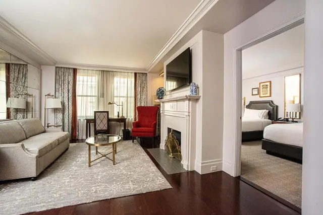 A living area with a grey sofa, a glass-topped coffee table on a patterned rug, a red armchair, a fireplace with a wall-mounted television, and a doorway leading to a bedroom with two beds.