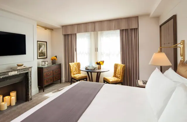 A bedroom with a king bed, a wall-mounted television, a marble fireplace, and a round table with two yellow armchairs by a window.