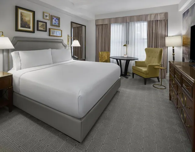 A king bed with white linens sits beside a yellow-green armchair, a round black table and a wooden dresser in a carpeted hotel room.