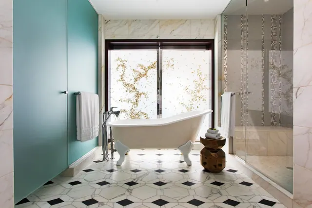 A freestanding clawfoot bathtub sits on a black-and-white marble tiled floor beside a glass-enclosed walk-in shower, with a frosted window behind.