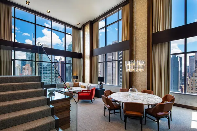 A two-storey living room with floor-to-ceiling windows overlooking city high-rises, a staircase, a sofa with red armchairs, and a marble dining table with leather chairs.
