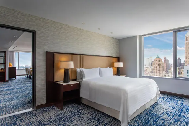 A bedroom with a king bed, bedside lamps and a large window overlooking city skyscrapers, with an open doorway to an adjoining dining area.