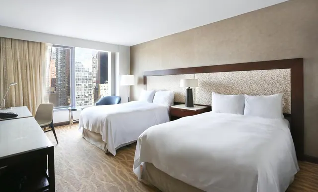 A hotel room with two beds made up with white bedding, a desk and armchair, and floor-to-ceiling windows looking out over city high-rises.