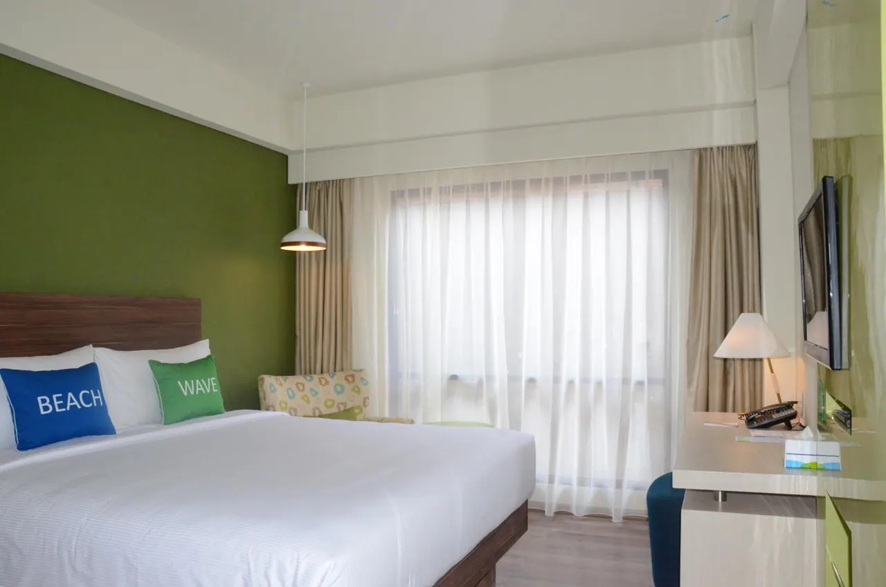 A bedroom with a made-up king bed, a green accent wall, decorative pillows, sheer curtains over a window, an armchair, a wall-mounted television and a work desk.