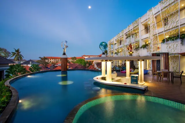 Dusk view of a large pool with a pool bar labelled Oasis, a tall sculpture, outdoor dining tables on the deck, and a multi-storey hotel building with a patterned lattice facade.