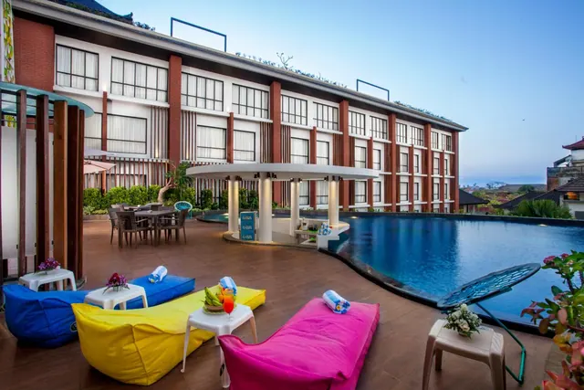 A large outdoor pool beside a timber deck with colourful bean bags, a small table holding fruit and a drink, a white circular pool bar, and a multi-storey hotel building behind.