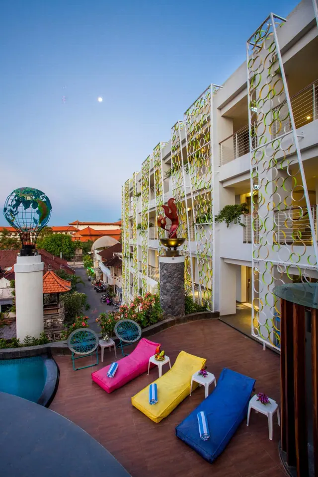 A terrace at dusk with pink, yellow and blue lounge cushions, white side tables and a pool edge in the foreground, with a street and a globe sculpture visible beyond.