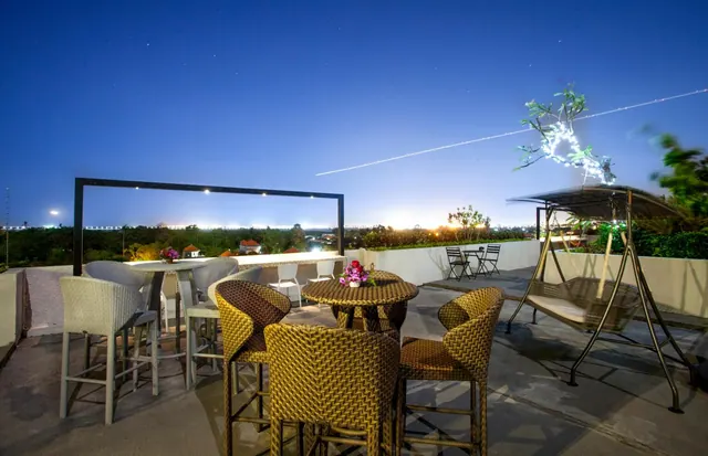 A rooftop terrace at dusk with wicker chairs and tables, a swing seat, and a view over distant trees and lights.