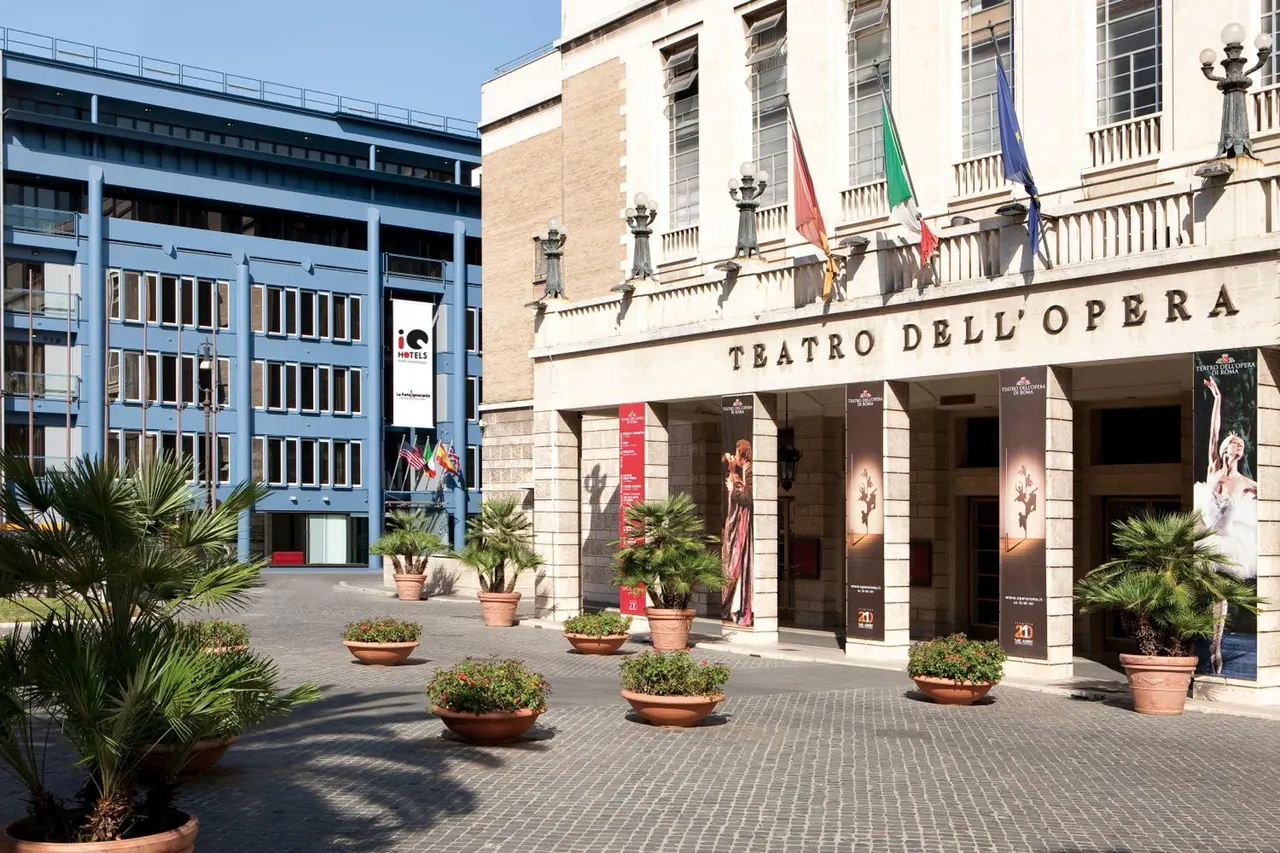 The facade of the Teatro dell'Opera stands across a paved square lined with potted plants, with a blue hotel building on the left.