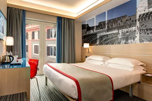A double bed with white bedding and a red-edged throw sits beside a desk with a kettle, under a large black-and-white Colosseum mural, with tall windows showing a street of buildings.