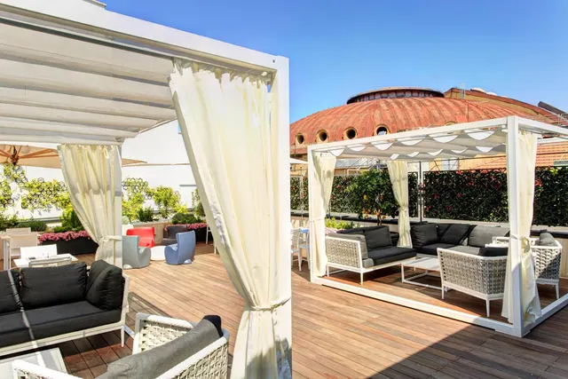 A rooftop terrace with white canopied cabanas, grey woven lounge sofas on a wooden deck, and a terracotta dome behind.