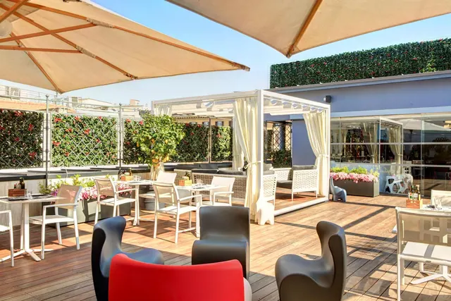 A rooftop terrace with wooden decking, cream parasols, white dining tables and chairs, dark armchairs in the foreground, and a white cabana with curtains.