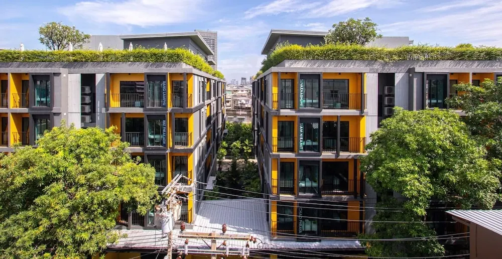 Exterior view of a multi-storey building with yellow and grey facades, balconies, rooftop greenery and trees, with a city street visible between two blocks.