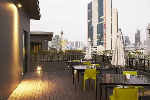 A rooftop terrace with dark tables, yellow chairs and closed white umbrellas overlooks high-rise buildings at dusk.
